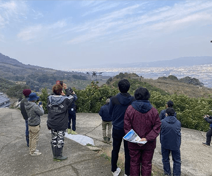 参加者がドローンの実演飛行を見学している様子。山と海を背景に、ドローンが上空を飛行している