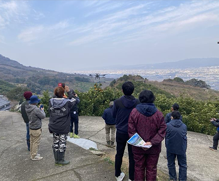 参加者がドローンの実演飛行を見学している様子。山と海を背景に、ドローンが上空を飛行している