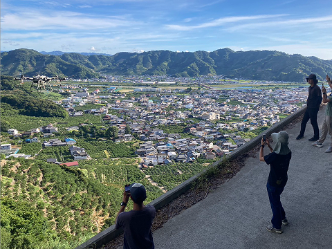 みかん畑と町並みを見下ろす高台から、参加者がドローンの飛行範囲を確認している様子